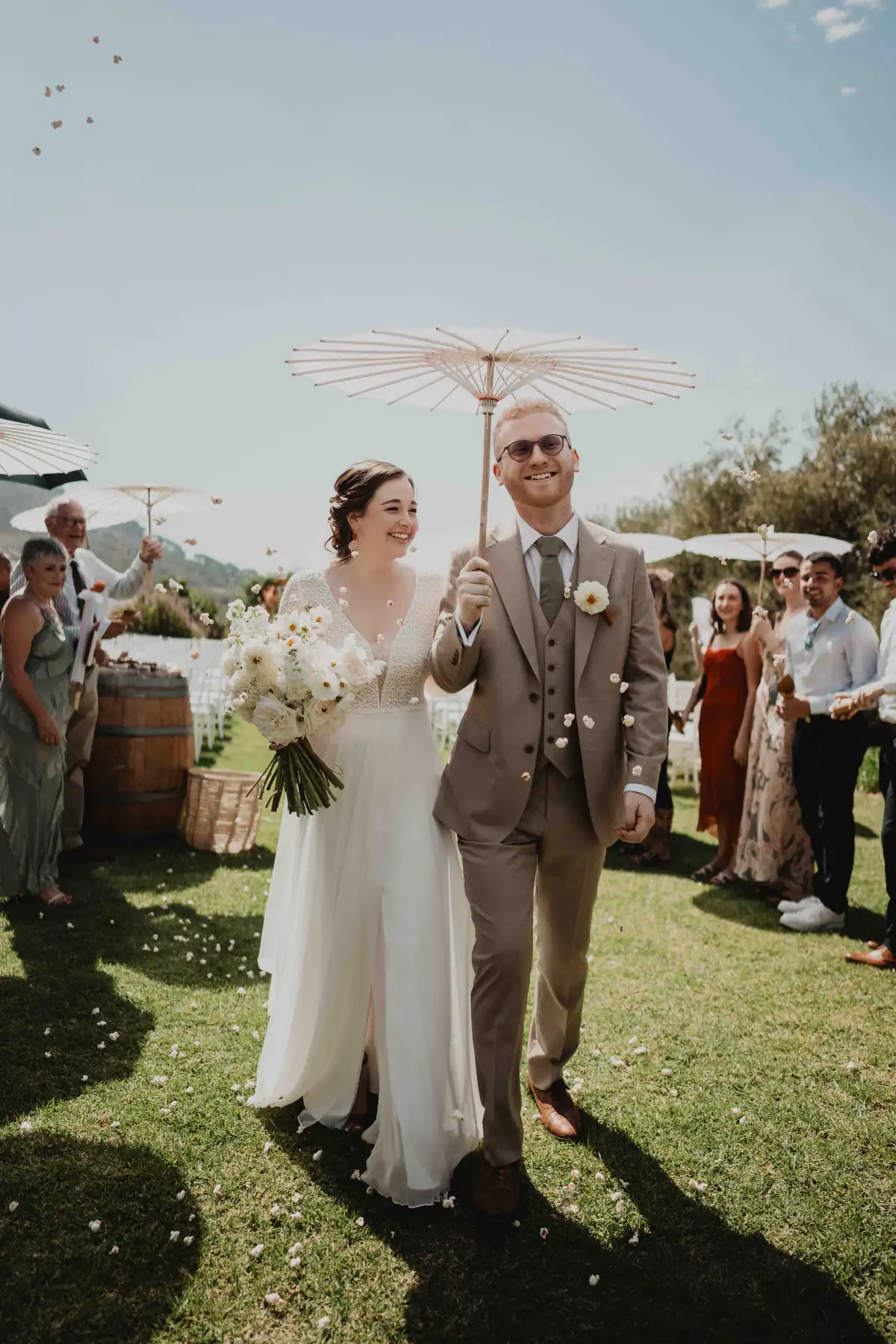 A bride and groom under a paper parasol, walking through confetti after their ceremony on the lawn