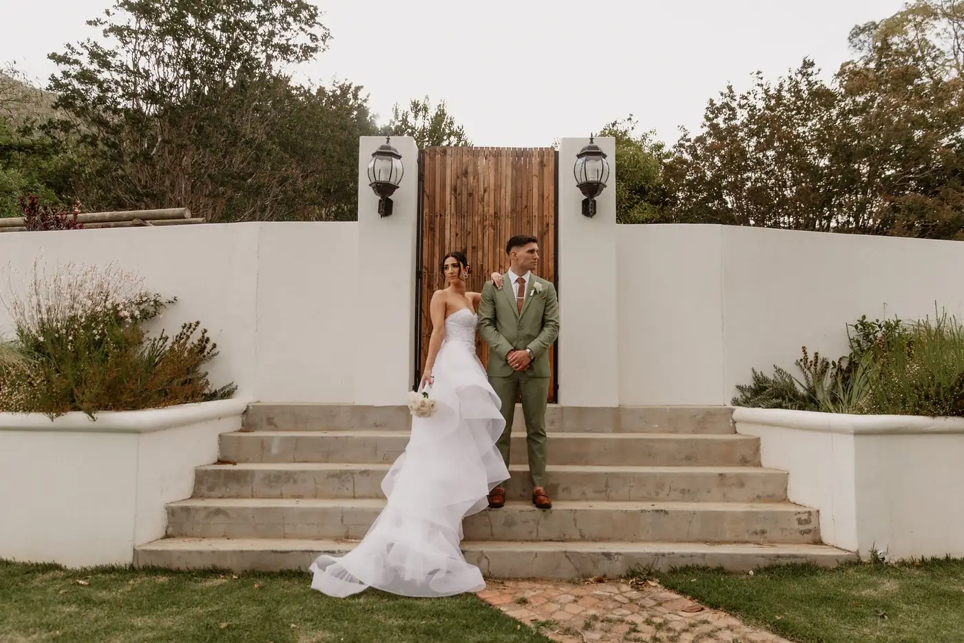 A bride and groom on the steps before a wooden gate in a white garden wall