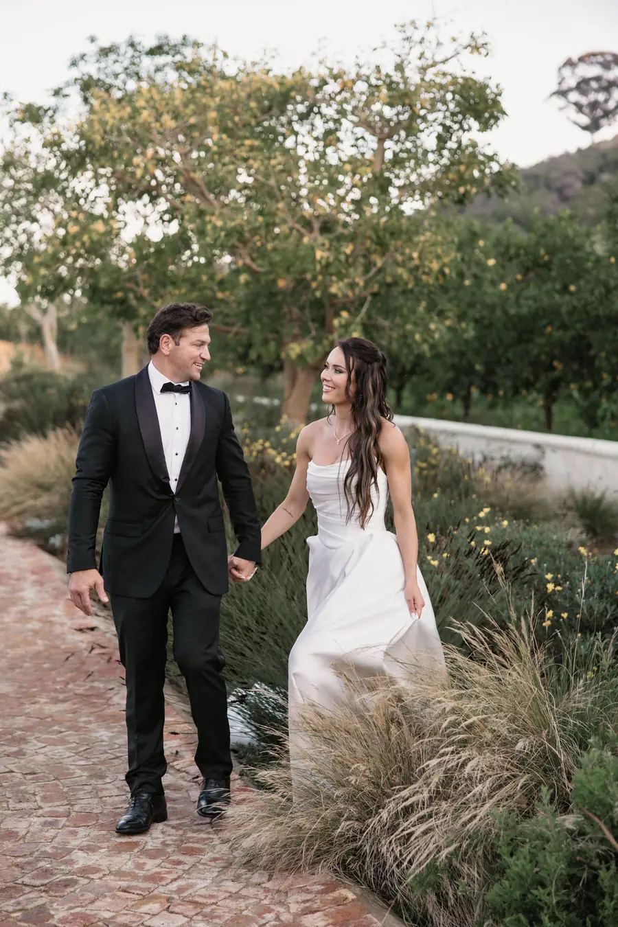 A bride and groom hand in hand on a brick path through the garden