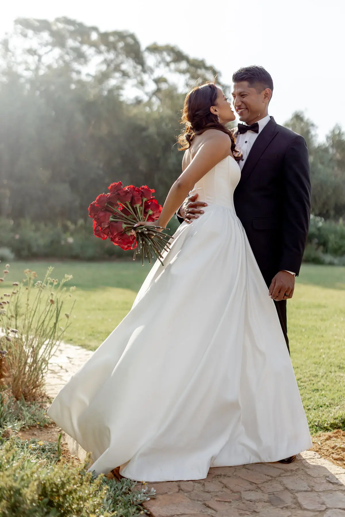 A smiling bride holding red roses, her groom’s arm around her, in the garden