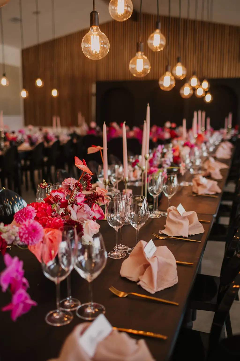 A long dinner table with pink and red flowers, tall candles and hanging bulbs