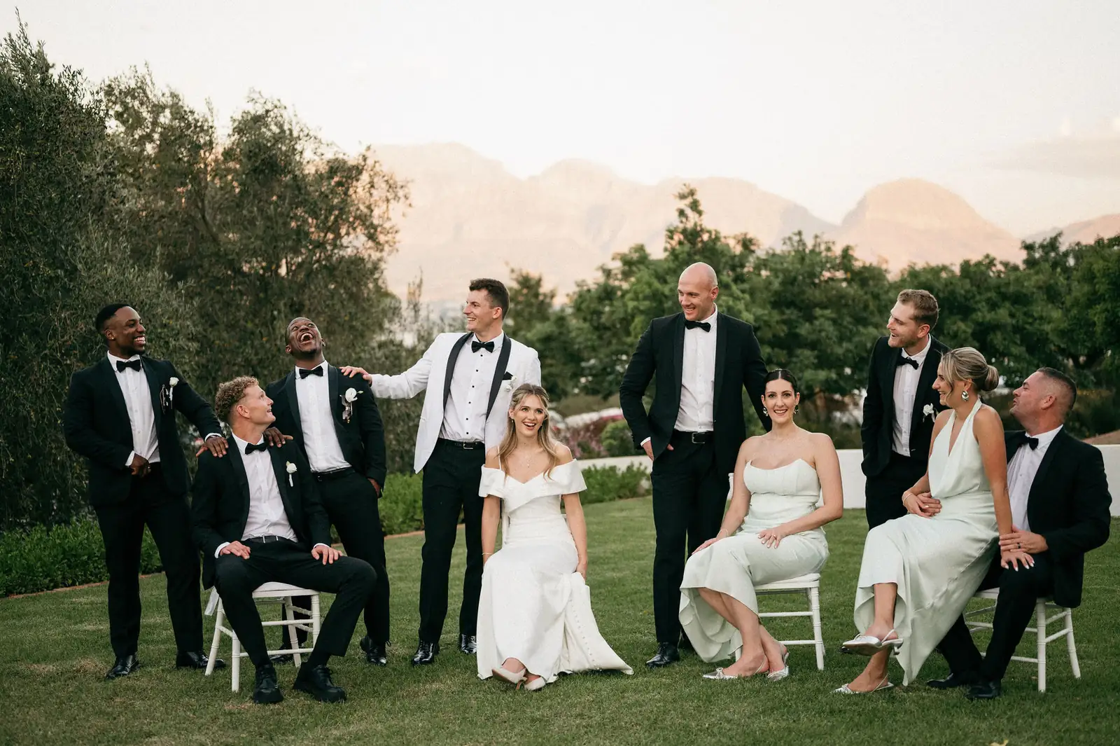 A bride, a groom and their wedding party laughing on the lawn, the mountains behind them
