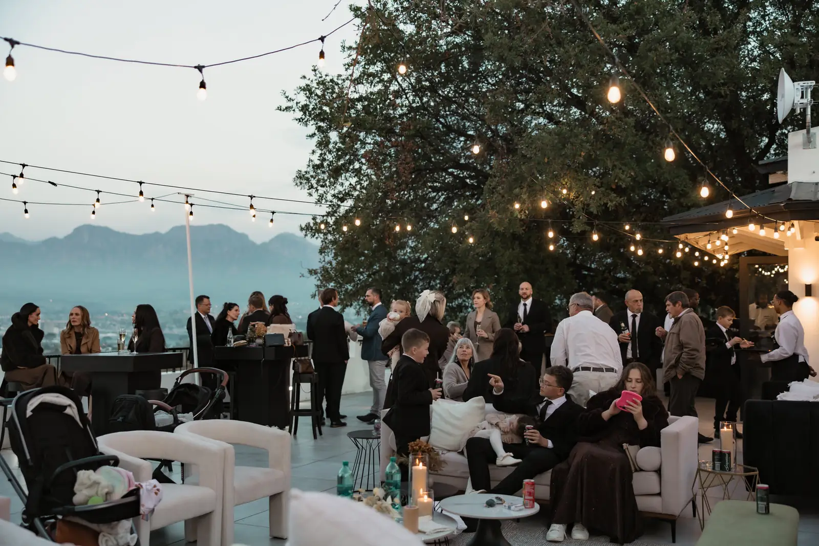 Guests in lounge corners on the patio at sunset, string lights overhead and the mountains beyond